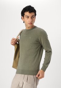 Zöld kötött pulóver kerek nyakkivágással, bordás mandzsettával és szegéllyel; világos bézs kordbársony nadrággal és hozzá illő táskával viselve.