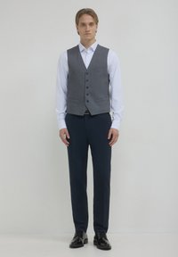 Junger Mann steht, trägt eine graue, geknöpfte Weste über einem weißen Hemd, eine marineblaue Hose und schwarze, polierte Lackschuhe vor einem einfarbigen hellen Hintergrund.