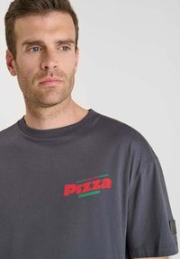 Man met lichte stoppels in een donkergrijs T-shirt met een klein rood en groen "Pizza" logo op de bovenste linkerborst, kijkt opzij.