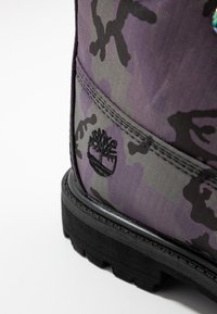 Bota de camuflaje morada con tejido texturizado, suela de goma negra y logo bordado. Costuras visibles y elementos de diseño distintivos.