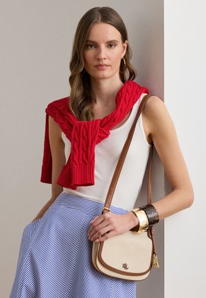 Donna con capelli lunghi che indossa una canotta bianca, maglione rosso appoggiato sulle spalle, gonna a righe blu, che tiene una borsa a tracolla beige e indossa braccialetti.