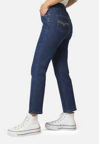 Jeans de mezclilla en azul oscuro con un diseño ajustado, que cuentan con un bolsillo trasero con costura decorativa, combinados con zapatillas altas blancas.