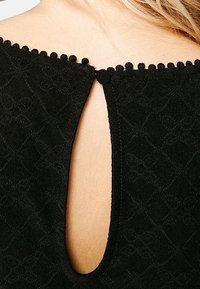 Prenda negra con un patrón texturizado y una pequeña abertura en la parte posterior, asegurada con un botón y un borde de pequeños pompones negros a lo largo del escote.