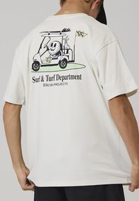 Maglietta in cotone bianco con grafica di un golf cart con un personaggio dei cartoni animati, testo "Surf & Turf Department" e accenti colorati.