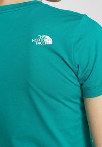 Blågrøn bomulds t-shirt med korte ærmer, med en hvid The North Face-logo på ryggen nær halsudskæringen. Glat tekstur, tætsiddende design.