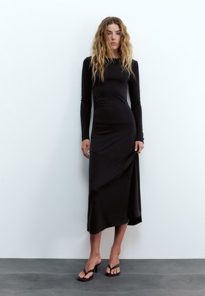 Kvinde med langt, bølget hår iført en tætsiddende sort midi-kjole med lange ærmer og sorte sandaletter med hæl, stående foran en hvid væg.
