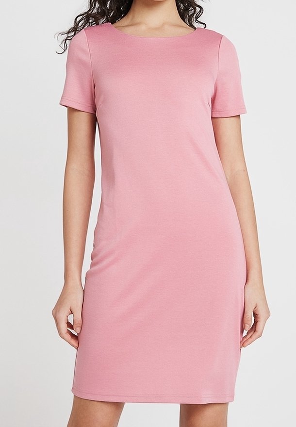 Femme portant une robe rose simple, à manches courtes, longueur genou, avec un col arrondi, debout devant un fond clair uni.