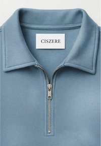 Helles blaues Poloshirt mit einem silbernen Halbreißverschluss und einem weißen rechteckigen Etikett mit der Aufschrift "CISZERE" innen am Kragen.
