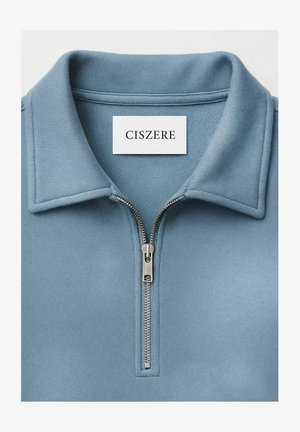 Lyseblå collared genser med en sølvfarget halvglidelås og hvit rektangulær etikett med teksten "CISZERE" inni kragen.