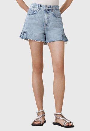 Short en jean bleu clair avec des ourlets effilochés et de petits détails à volants, porté avec des sandales plates argentées et un top blanc rentré.