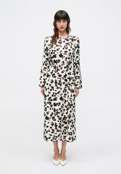 Long-sleeve white dress with black and brown leopard print, fitted at the waist, flowing skirt, worn with cream-colored pointed heels.