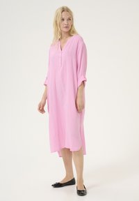 Rozā garā tunika kleita, izgatavota no viegla materiāla, ar V veida kaklu, pogu rindu, sarullētām piedurknēm un sānu šķeltnēm, lai nodrošinātu ērtu kustību.