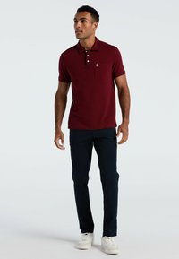 Bordeauxrotes Poloshirt mit Brusttasche, kontrastierendem schwarzen Kragen und dreiknöpfiger Knopfleiste. Kombiniert mit navyblauen Cargo-Hosen und weißen Sneakers.
