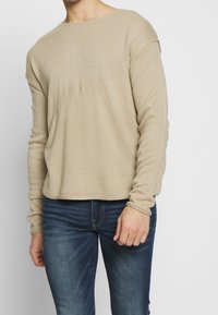 Beżowy, długi sweter z dzianiny z okrągłym dekoltem i prążkowaną fakturą, zestawiony z niebieskimi jeansami. Casualowy krój z subtelnym przeszyciem.