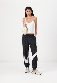 Top cinza, calças de treino pretas com padrão de swoosh branco, mala de ombro bege e ténis de cor clara. Design simples e desportivo.