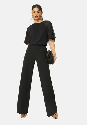 Kvinde iført en sort gennemsigtig kortærmet bluse, højtaljede bukser med vide ben, holder en sort struktureret clutch og har snøresandaler på.