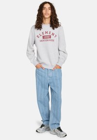 Sudadera gris con gráfico rojo "ELEMENT 1992 SKATEBOARDS", combinada con jeans azul claro con un patrón texturizado y zapatillas grises.
