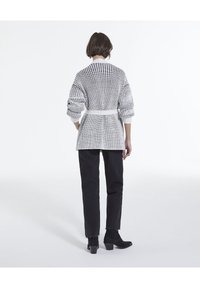 Kvinna med kort hår iförd en vit och svart stickad cardigan med bälte, svarta byxor och svarta klackiga ankelstövlar, vänd bort.