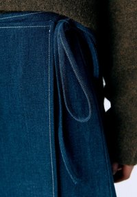 Denimforklæde i mørkeblå farve med sidesnøring, hvide syninger og en glat overflade, båret over et brunt stof.
