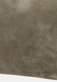 Zachte grijze suède oppervlakte met kleine, gecentreerde metalen tekstlogo "COCCINELLE" reliëf aangebracht nabij de onderrand.