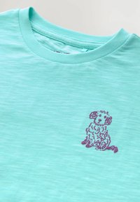 Camiseta de manga corta verde menta con una textura en la tela, que presenta un diseño de perro bordado en púrpura en el lado izquierdo del pecho.