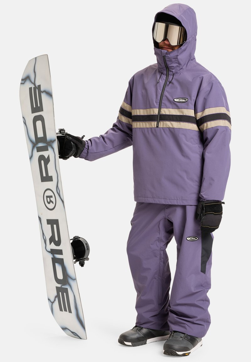 Giacca e pantaloni da snowboard viola con strisce nere e beige; persona che tiene una tavola da snowboard bianca con grafiche e legami neri.