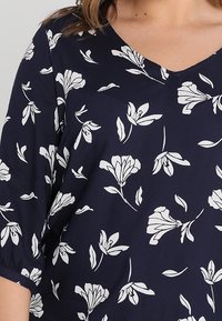 Laivastonsininen pusero, jossa on V-pääntie ja valkoisia kukkakuviota. Materiaali näyttää sileältä, ja siinä on lyhyet, väljät hihat sekä rento istuvuus.