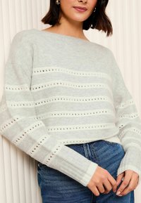 Pullover gri deschis cu dungi orizontale albe, având modele din dantelă. Textură moale, mansete largi și decolteu rotunjit. Asortat cu blugi.