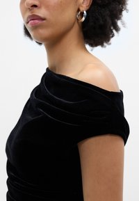 Μαύρο βελούδινο φόρεμα με ντραπέ λαιμόκοψη, εκτός ώμου σχέδιο και κοντό μανίκι. Περιλαμβάνει λαμπερά ασημένια σκουλαρίκια σε σχήμα κρίκου ως αξεσ�ουάρ.