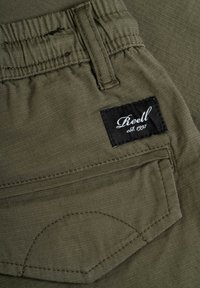 Tejido verde oliva con textura suave; cinturilla elástica; presenta una etiqueta negra con "Reell est. 1997" y detalle de costura en el bolsillo.