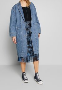 Žena v oversized modrém denimu kabátu, černém topu, modré vzorované plisované sukni a námořnických vysokých teniskách, stojící na šedém pozadí.