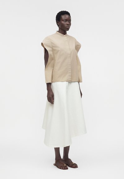 Γυναίκα με μπεζ αμάνικο πουκάμισο με κουμπιά, λευκή μίντι φούστα και καφέ επίπεδες σανδάλια, στέκεται μπροστά σε απλό λευκό φόντο.