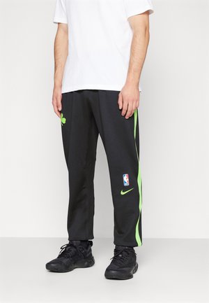 Mężczyzna noszący czarne spodnie dresowe Nike z zielonymi paskami i logo NBA, w połączeniu z czarnymi butami sportowymi i białą koszulką z krótkim rękawem.