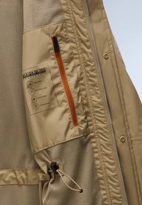 Μπεζ σακάκι με επένδυση από δίχτυ, που διαθέτει πορτοκαλί τσεπάκι με φερμουάρ, κορδόνι στη μέση και ετικέτα που υποδεικνύει ύφασμα ανθεκτικό στο νερό και διαπνέον.