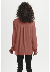 Rustfarbenes langärmliges Blusenshirt aus einem strukturierten, zerknitterten Stoff. Verfügt über eine Passe mit Raffungen im Rücken und einen abgerundeten Saum. Getragen mit schwarzen Hosen.