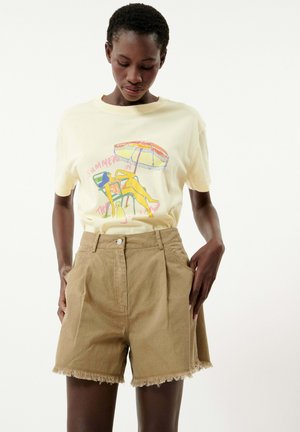 T-shirt jaune à manches courtes avec un motif coloré sur le thème de la plage ; associé à un short en denim beige taille haute avec un ourlet effiloché et des poches latérales.