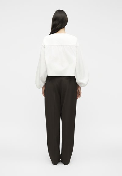 Γυναίκα που στέκεται με την πλάτη, φοράει λευκό πουκάμισο με μακριά μανίκια και φουσκωτά μανίκια και ψηλόμεση, φαρδιά μαύρη παντελόνα.