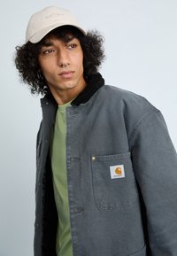 Graue Canvasjacke mit Kordkragen, goldenen Druckknöpfen und Fronttasche. Getragen über einem grünen Hemd, mit einer beigen Kappe.