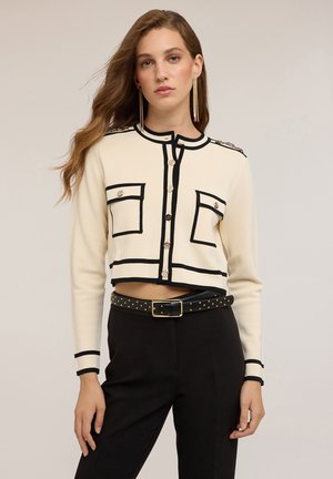Cardigan corto crema con bordi neri, due tasche frontali e bottoni dorati. Abbinato a pantaloni neri e una cintura con borchie. Maniche lunghe.