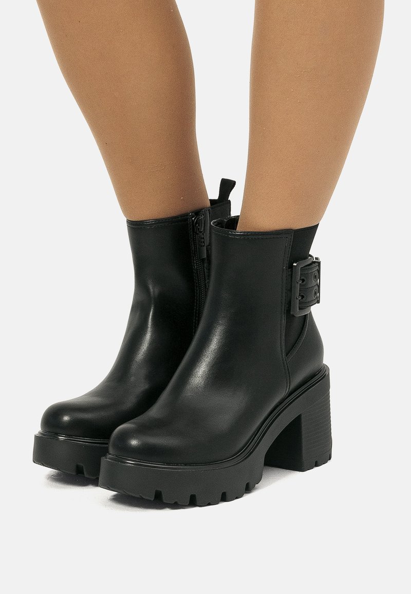 Stivali neri in pelle fino alla caviglia con tacco spesso, cerniera laterale e dettaglio con fibbia, indossati con collant trasparenti color carne.