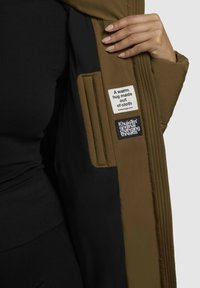 Interno di una giacca isolante marrone con rivestimento nero, dotato di due etichette: una con testo e una con logo. Texture liscia e imbottita.