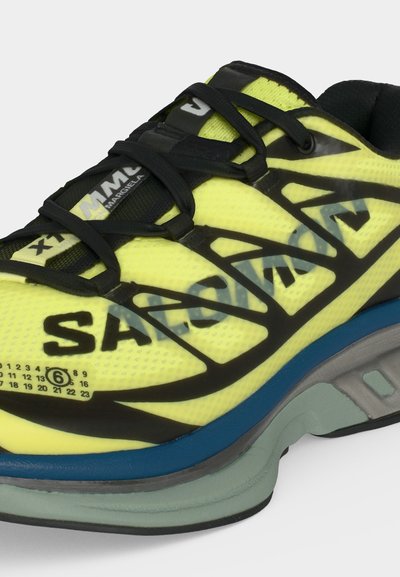 Αθλητικό παπούτσι νέον κίτρινο και μαύρο με το κείμενο "SALEMM", μαύρα κορδόνια, μπλε και γκρι σόλα, και μικρό αριθμητικό σχέδιο κοντά στην περιοχή των δακτύλων.
