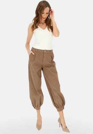 Kobieta w białym topie bez rękawów, brązowych krótkich spodniach z wysokim stanem i marszczonymi mankietami oraz beżowych szpilkach, stojąca z jedną ręką w kieszeni.