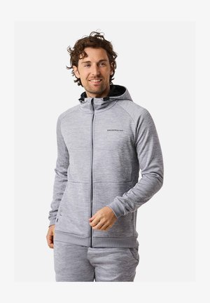 Muž s kučeravými vlasmi oblečený v svetlosivú mikinu na zips a zodpovedajúce tepláky, usmievajúci sa a pozerajúci sa do strany.