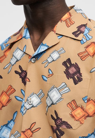 Chemise à manches courtes beige avec des motifs colorés de lapins robots en bleu, orange, blanc et marron, dotée d'une poche poitrine et de boutons.