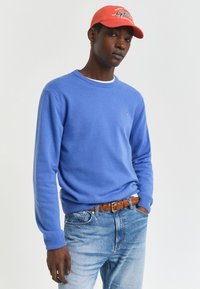 Junger Mann in einem blauen Pullover, hellblauen Jeans, braunem geflochtenen Gürtel und einer roten Baseballkappe vor einem einfarbigen Hintergrund.