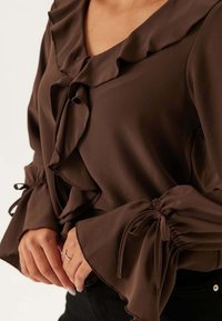 Blusa marrone con volant, scollo a V e maniche svasate, con lacci arricciati ai polsini, realizzata in tessuto liscio e leggero.