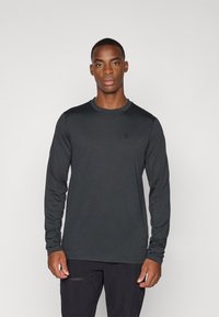 Dunkelgraues Langarmshirt mit Rundhalsausschnitt, glatter Textur und kleinem Logo auf der Brust, getragen mit schwarzen Hosen.
