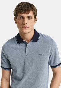 Lichtblauwe poloshirt met marineblauwe kraag, voorzien van een subtiele textuurpatroon en een klein "GANT" logo op de borst. Korte mouwen, klassieke pasvorm.