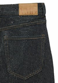 Tamni traperice od denima s teksturiranim uzorkom malih zakovica, sa kožnom oznakom označenom "VMD" i kontrastnim narančastim šavovima na džepovima.